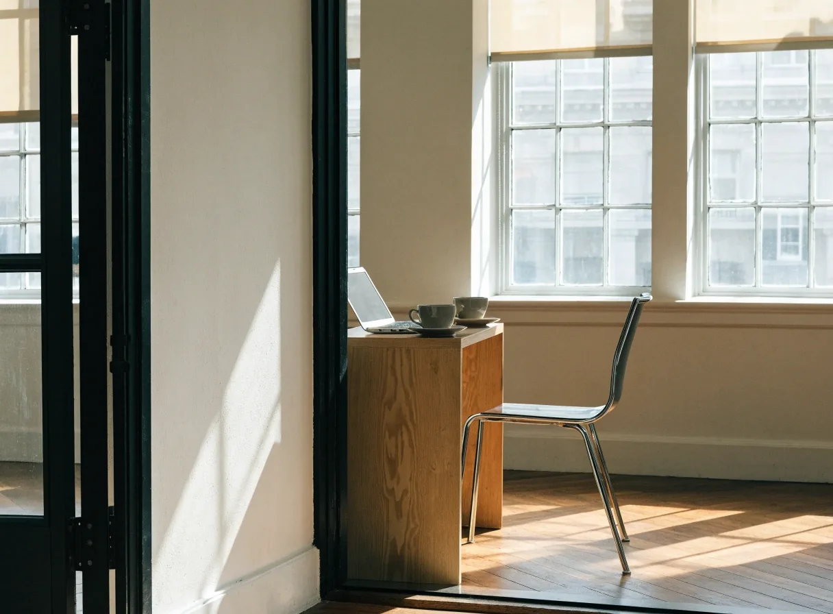Light consultation table with an open notebook, closed laptop and empty chair across, lit by soft lateral window daylight